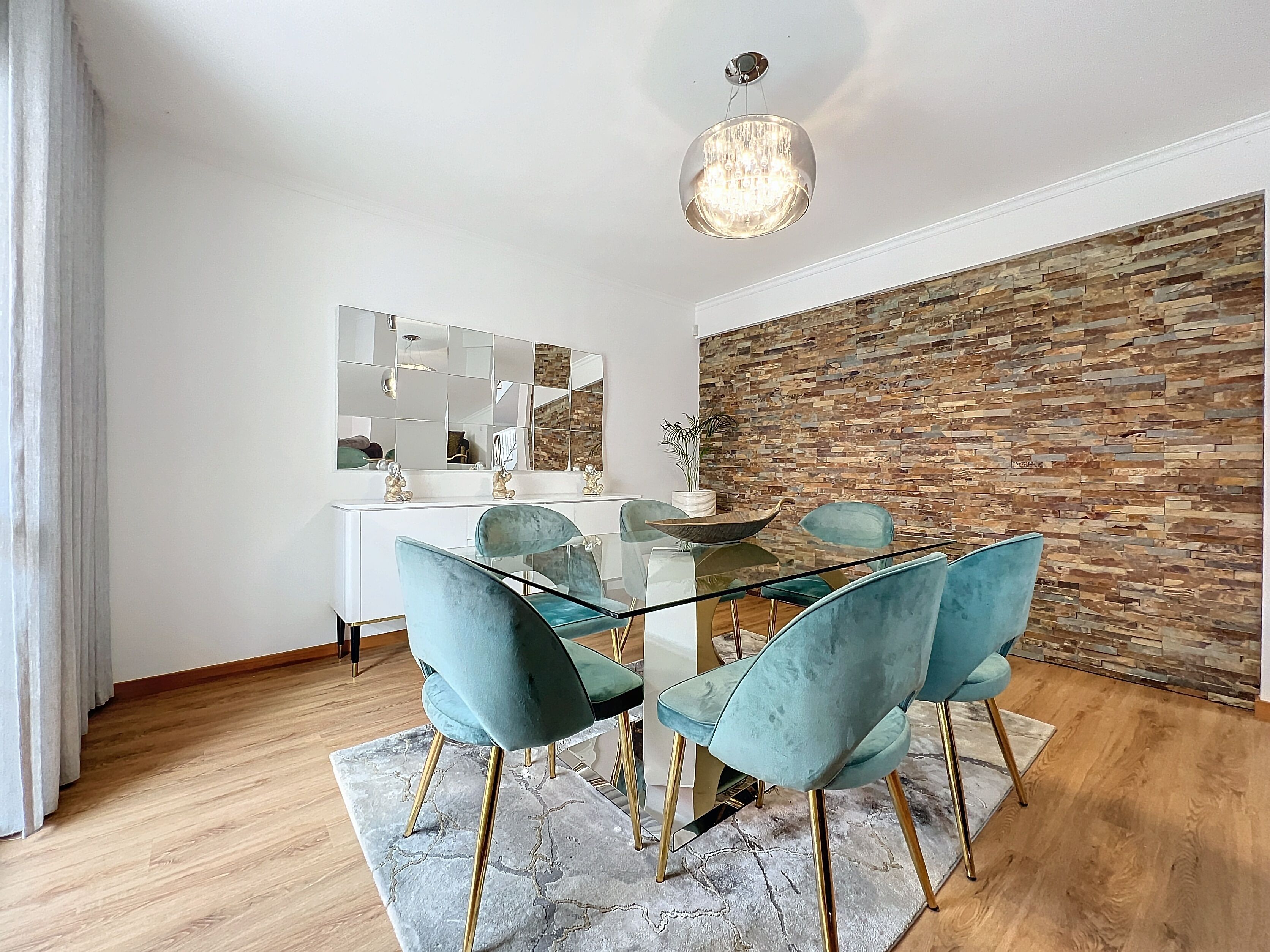 Un espace repas moderne avec une table en verre entourée de chaises turquoise, un meuble console blanc et un mur en pierre.