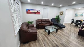 Una sala de estar con un sofá de cuero marrón, una mesa de centro de vidrio y un sillón negro. Hay una zona de comedor con una mesa y sillas, y un cuadro en la pared.