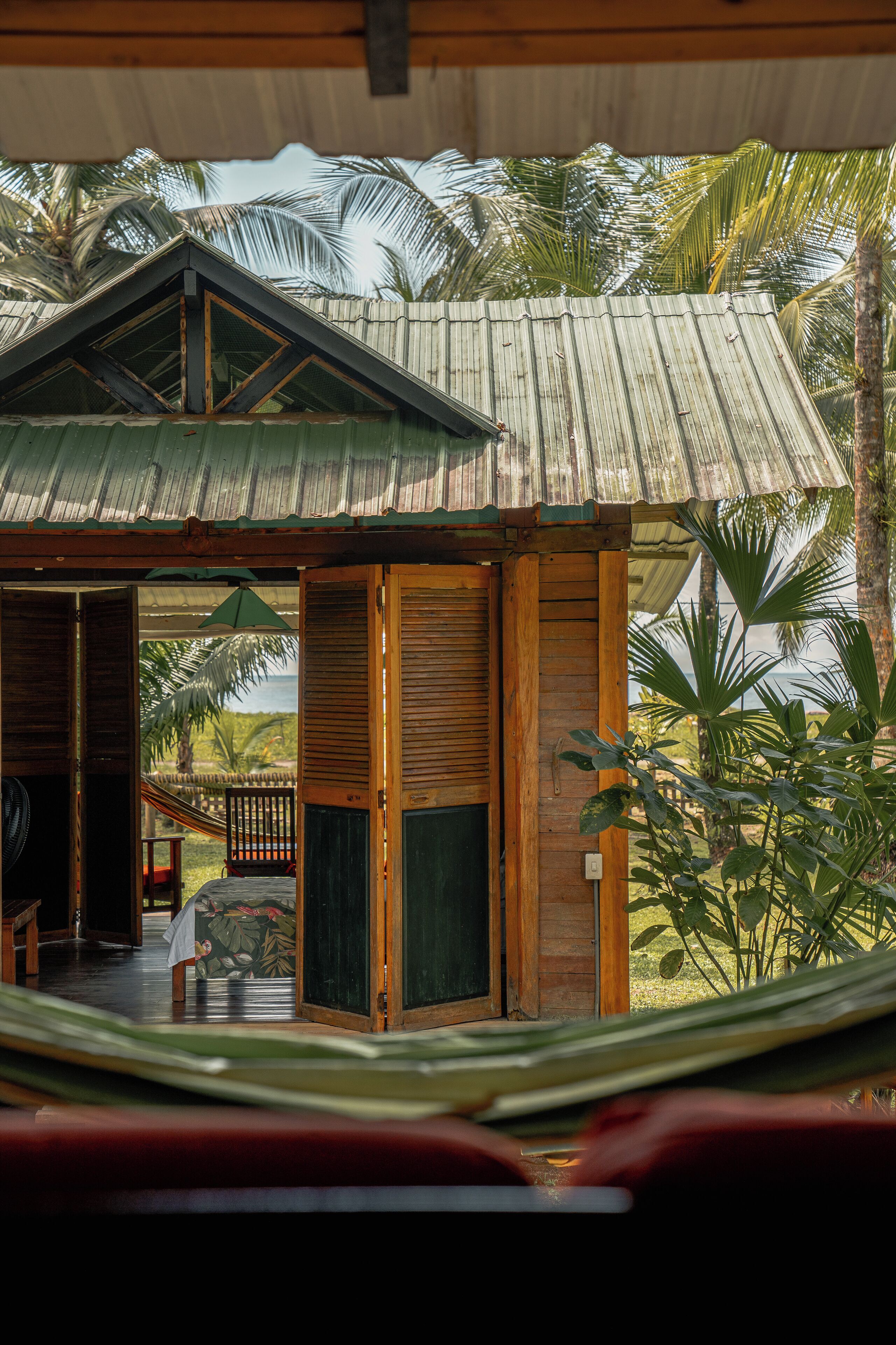 A wooden structure with a corrugated metal roof, a hammock, and a view of palm trees.