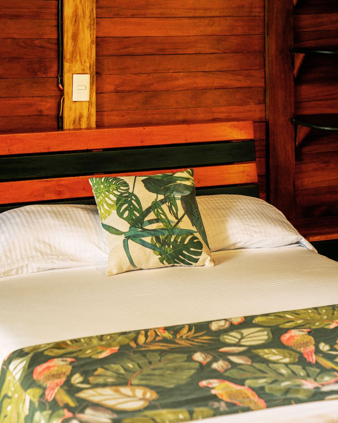Un lit avec une tête de lit en bois, un oreiller blanc et un coussin décoratif orné d’un motif de feuilles tropicales.
