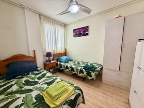 Une chambre avec deux lits, un ventilateur de plafond, une fenêtre avec des rideaux, une table de chevet avec une lampe et une armoire.