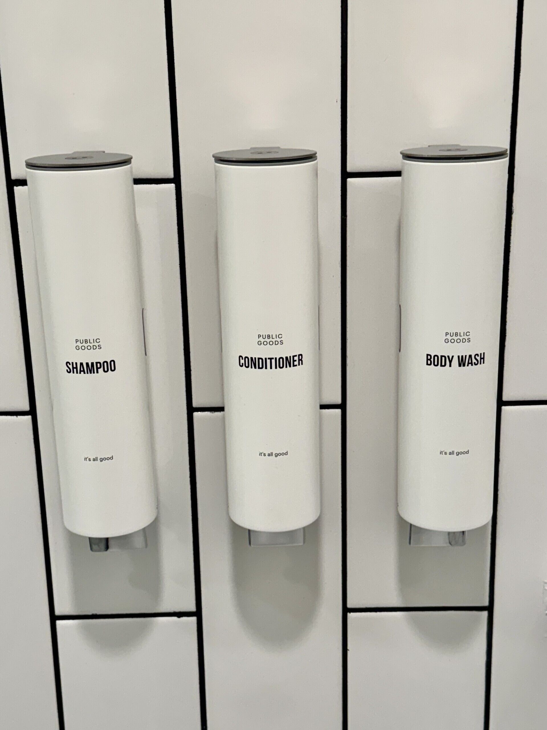 Three white dispensers labeled 'Public Goods' for shampoo, conditioner, and body wash, each with a lid and a pump.