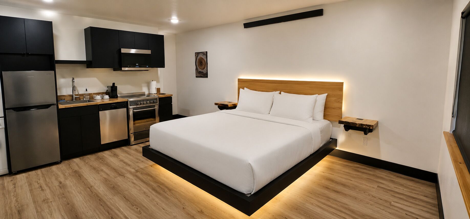 A modern hotel room with a bed, a kitchenette, and a wooden floor.