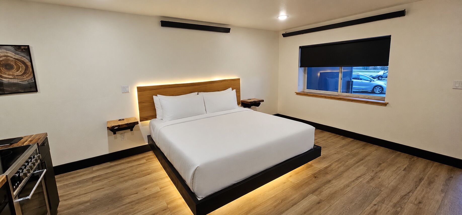 Een moderne hotelkamer met een bed, nachtkastje, en een raam met uitzicht op geparkeerde auto's.
