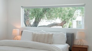 Une chambre avec une grande fenêtre, un lit recouvert de draps blancs, deux tables de chevet avec des lampes, et une vue sur les arbres à l’extérieur.