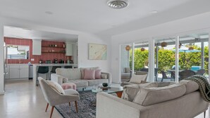 Un salon moderne avec un canapé, des fauteuils et une table basse. On aperçoit une cuisine au fond, avec des carreaux rouges, et un coin repas avec des sièges d’extérieur.