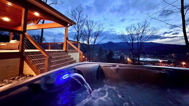 Un jacuzzi iluminado en azul, con una terraza de madera y vistas a montañas y un pueblo al fondo.