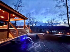A hot tub with a blue light, wooden deck, and a view of mountains and a town in the background. - Glamp House by Hiawassee Glamping (Hiawassee)