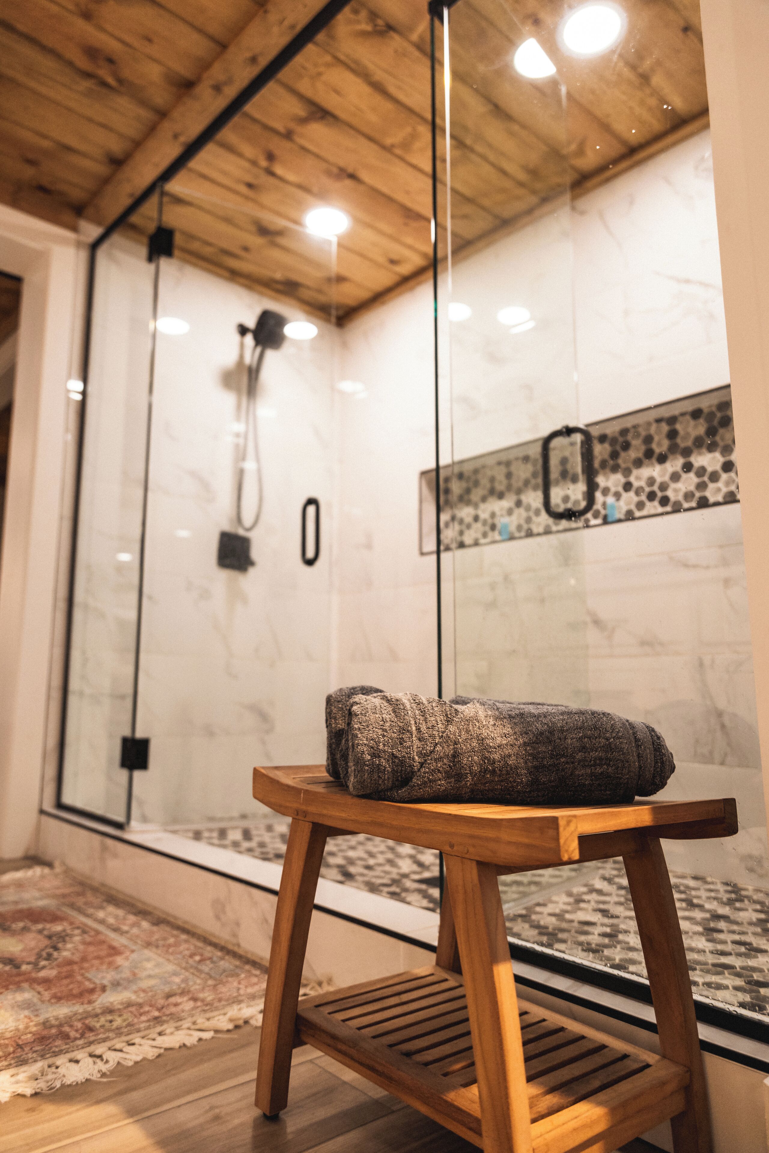 A bathroom with a glass shower enclosure, wooden floor, and a small wooden table with a towel.