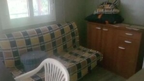 A room with a plaid sofa, a white plastic chair, a wooden cabinet, and a window with blinds. - Mobile home included in a complex of villas with swimming pool (Il Poggio)