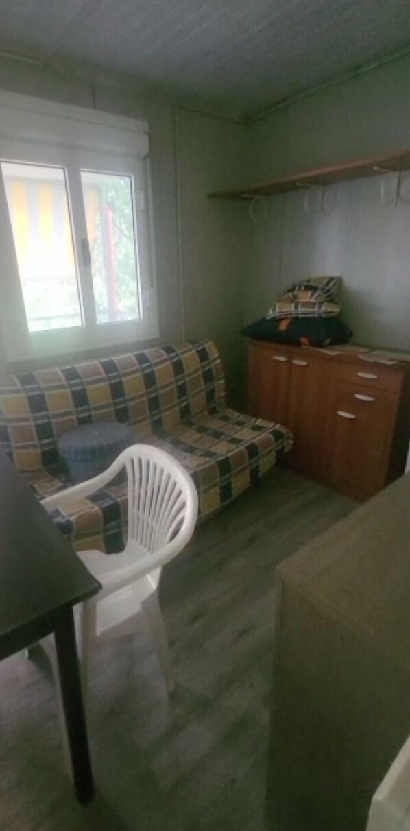 Une pièce avec un canapé à carreaux, une chaise en plastique blanche, un meuble en bois et une fenêtre avec des stores.