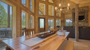 A wooden cabin interior with a dining table, chairs, a fireplace, and large windows.