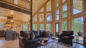 A spacious living area with a large wooden ceiling, a dark leather sofa, a coffee table, and a view of the outdoors through large windows. - Birch Lodge by Avantstay Indoor Pool, Theater, Game Room & Stunning Views! (Gatlinburg)