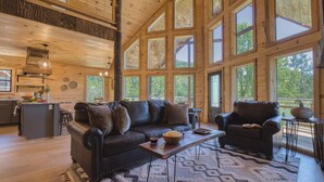 A spacious living area with a large wooden ceiling, a black leather sofa, a wooden coffee table, and a view of the outdoors through large windows. - Birch Lodge by Avantstay Indoor Pool, Theater, Game Room & Stunning Views! (Gatlinburg)
