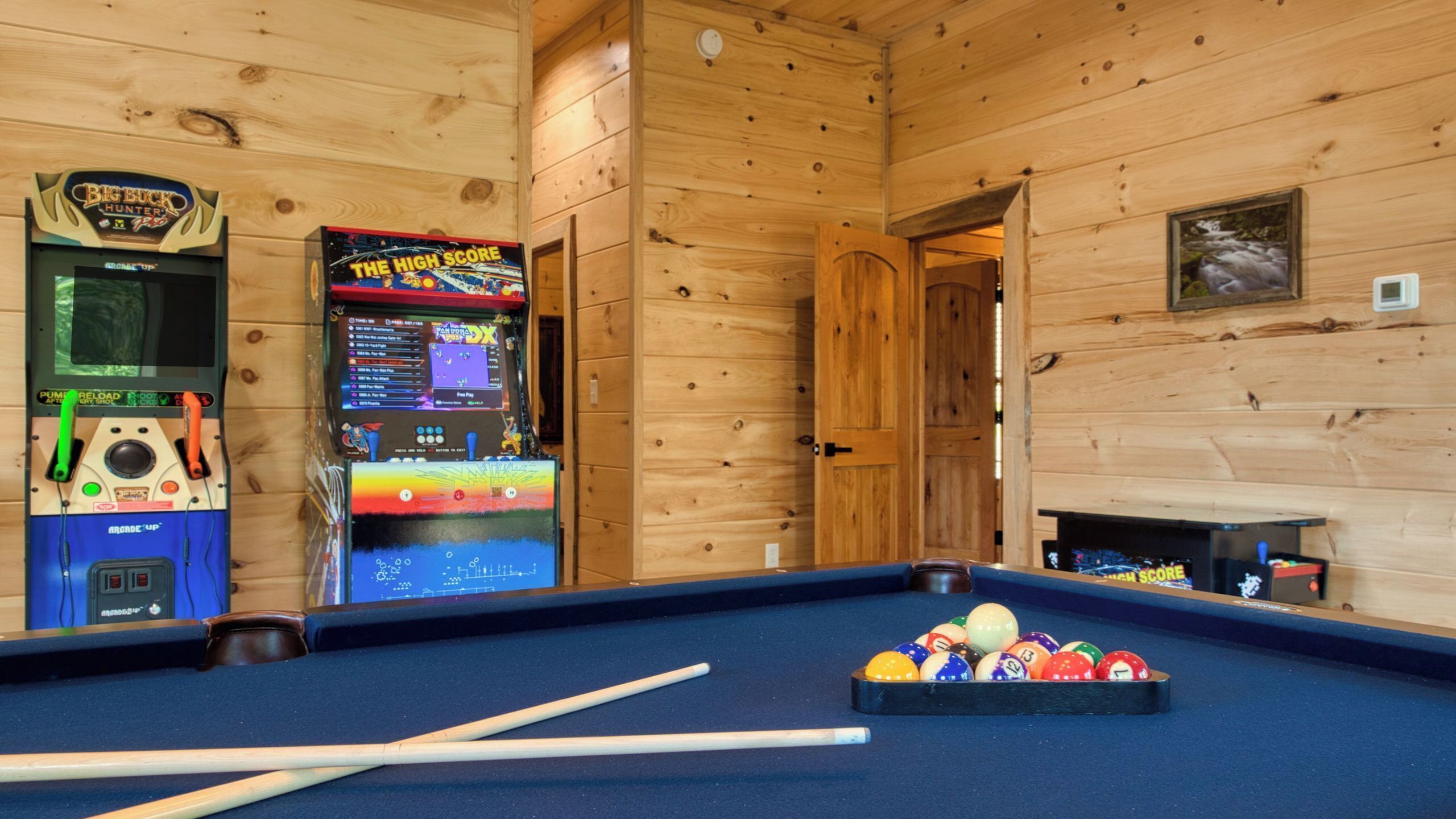 A pool table with billiard balls and cues, arcade machines, and wooden walls.