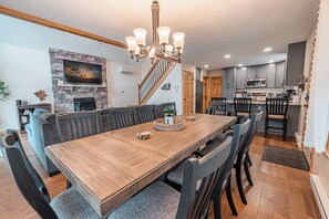 A spacious dining area with a wooden table and chairs, a stone fireplace, and a modern kitchen in the background. - NEW Large Group Summer Getaway W/hot Tub + Game Room! (Albrightsville)