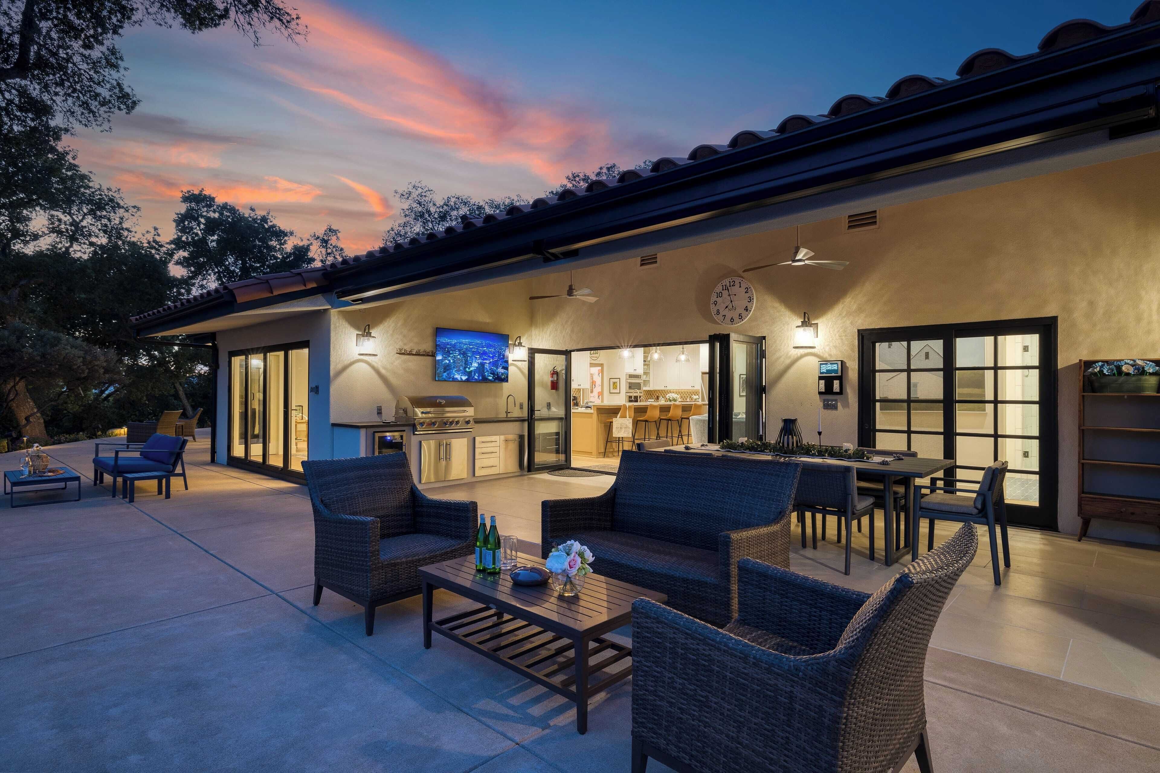 An outdoor patio area with wicker furniture, a dining table, and a kitchen visible through glass doors.