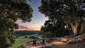 A circular outdoor seating area with chairs and a table, surrounded by rocks and trees, overlooking a vineyard and hilly landscape. - Rancho Robles by Avantstay Vineyard Villa w/ Views, Pool & Privacy (Paso Robles)
