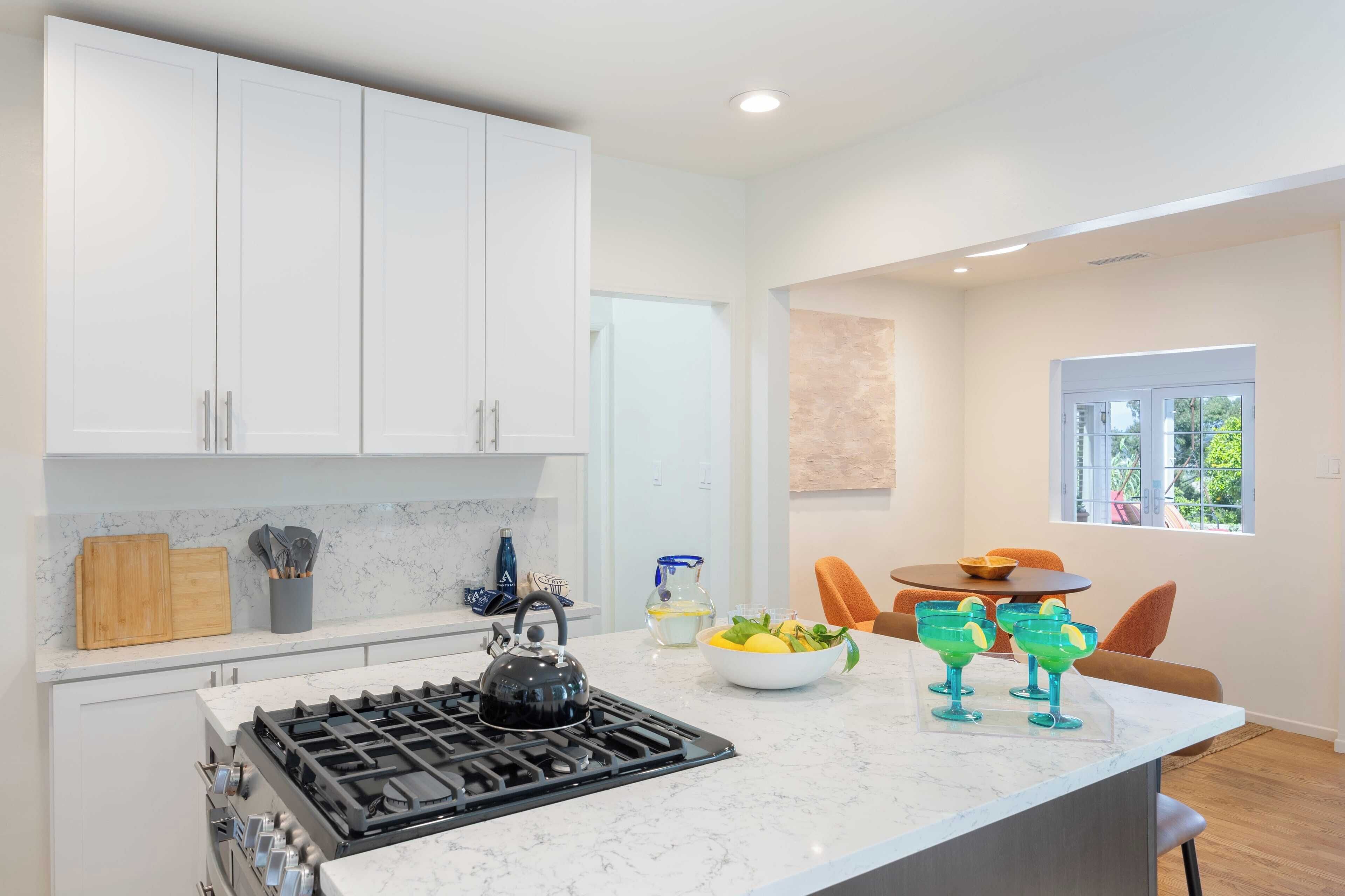 A modern kitchen with white cabinets, a gas stove, and a marble countertop. There is a bowl of fruit and green glasses on the counter.