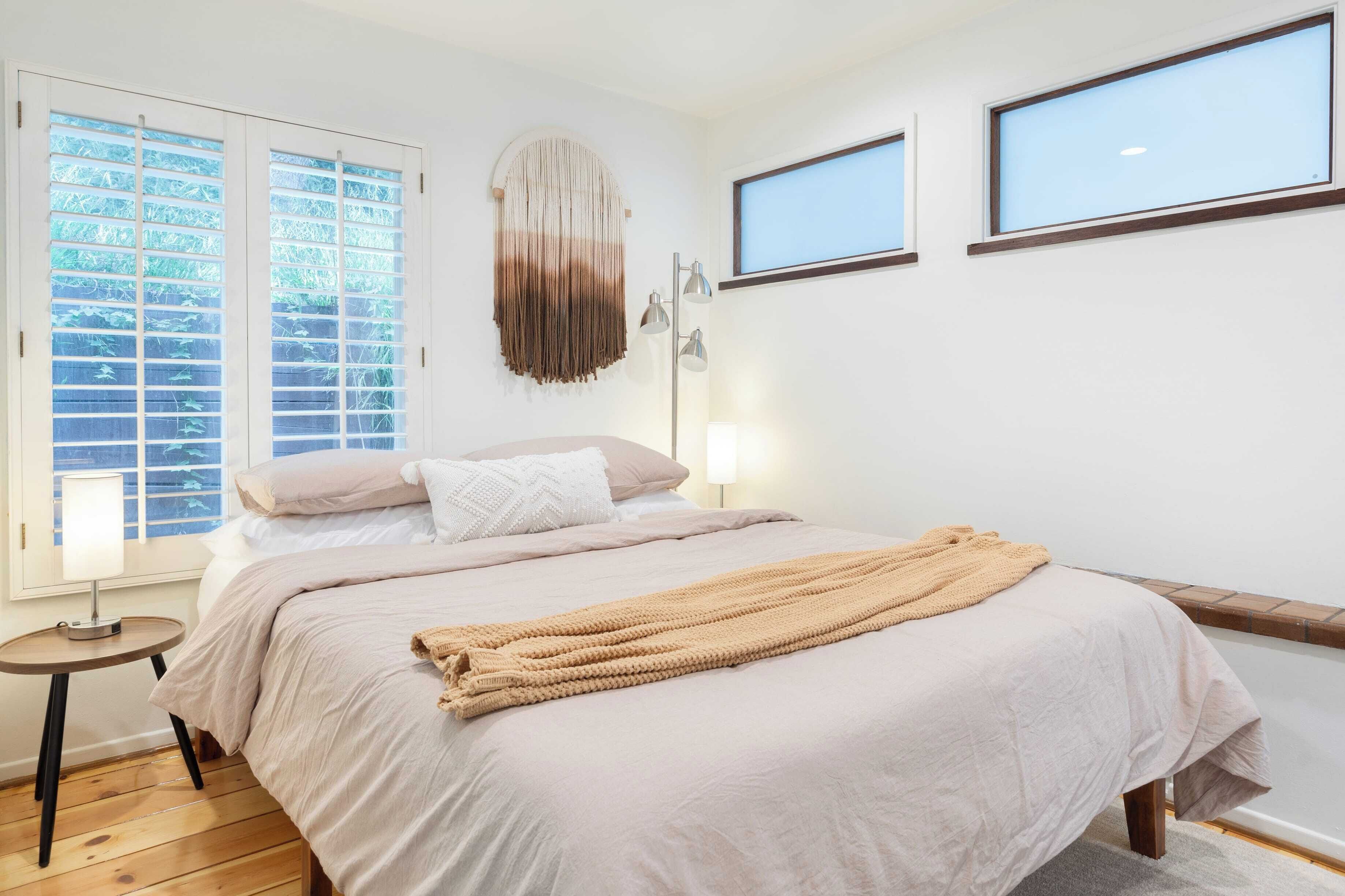 Un dormitorio con cama, lámpara de noche, luz en la pared y ventana con persianas.