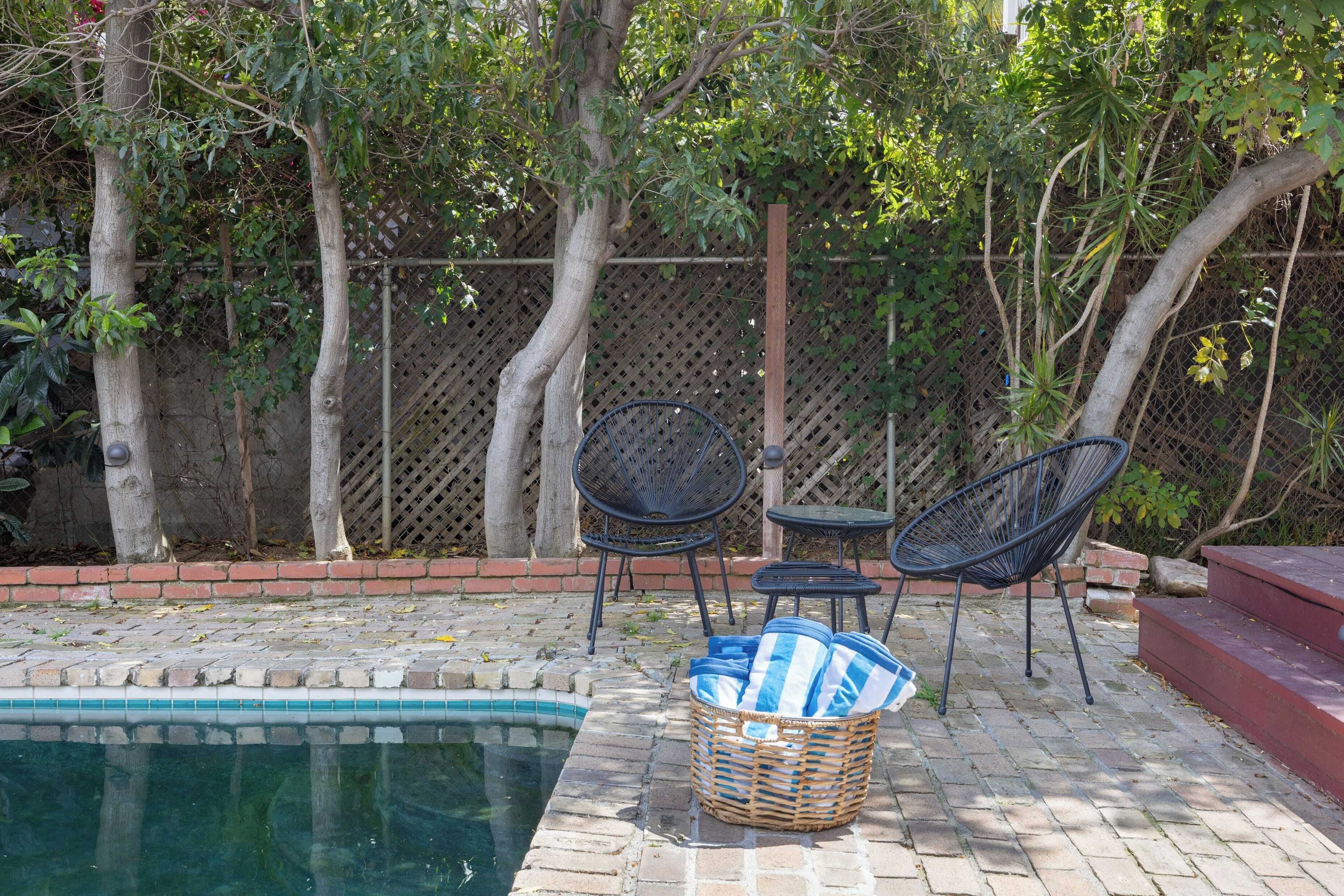 A poolside area with a brick patio, two black metal chairs, a small table, and a wicker basket with items.