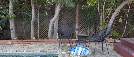 A poolside area with a brick patio, two black metal chairs, a small table, and a wicker basket with items.
