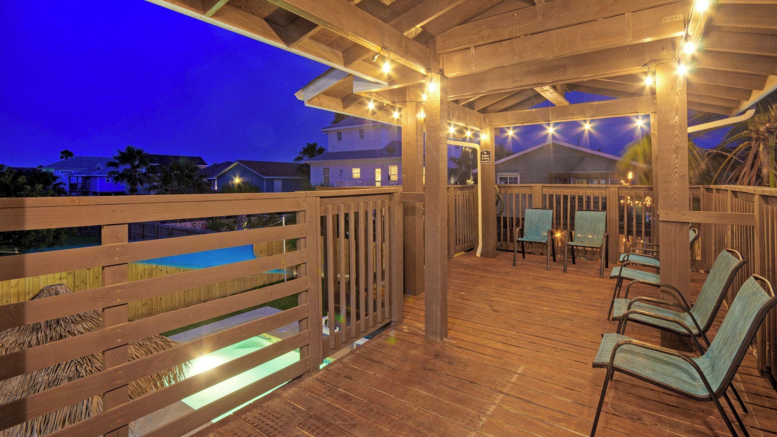 Une terrasse en bois, dotée de chaises et de guirlandes lumineuses, qui donne sur une piscine.
