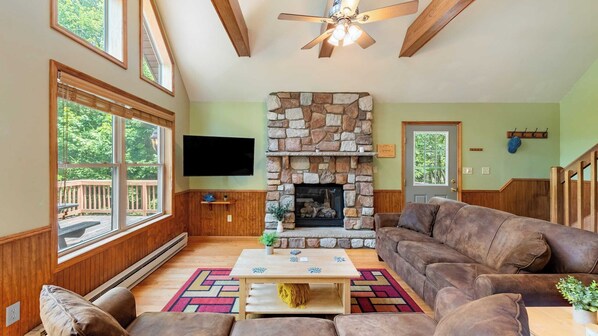 A cozy living room with a stone fireplace, wooden furniture, and large windows.