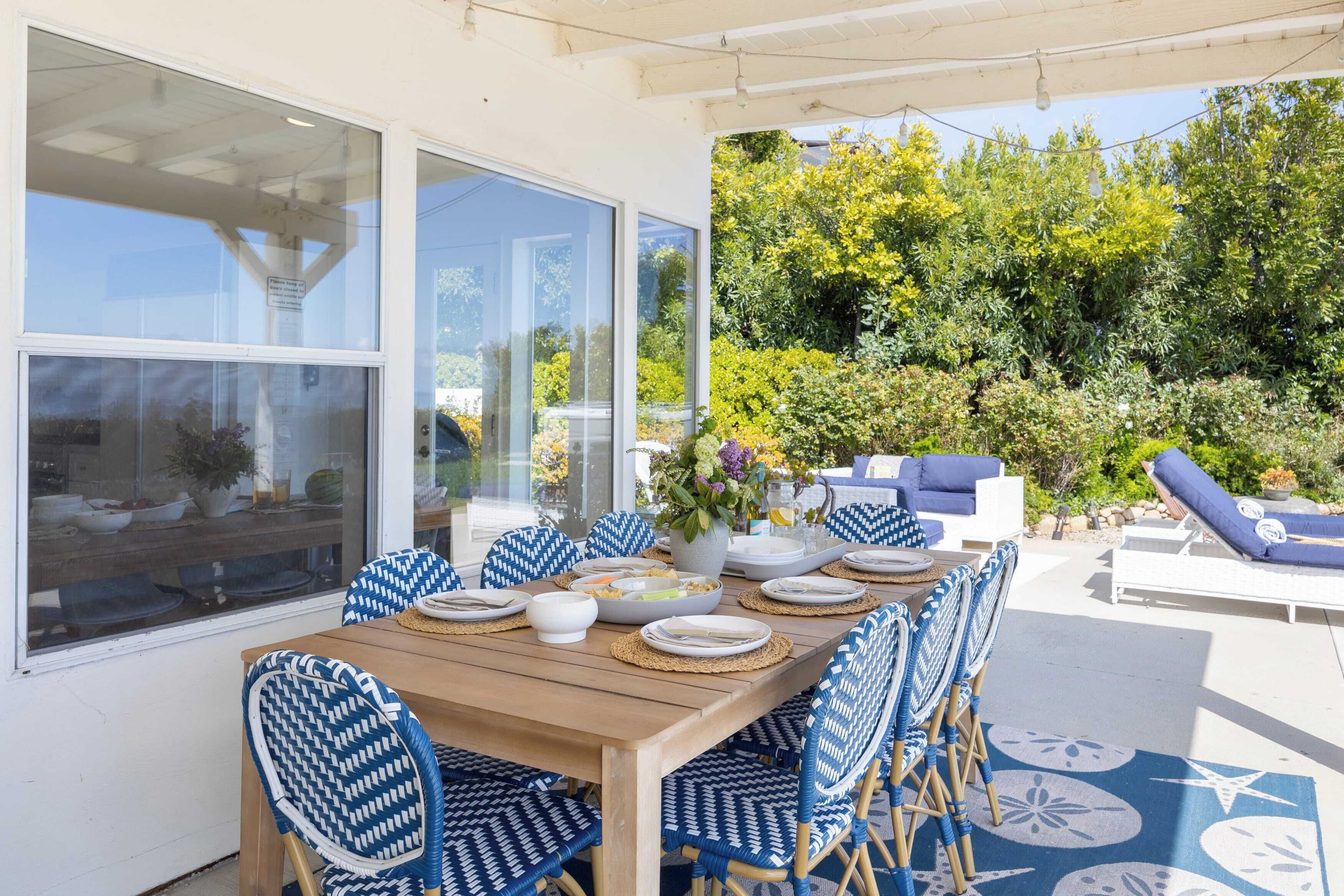 Un área de comedor al aire libre techada con una mesa de madera dispuesta para cuatro personas, rodeada de sillas con estampado azul y blanco.