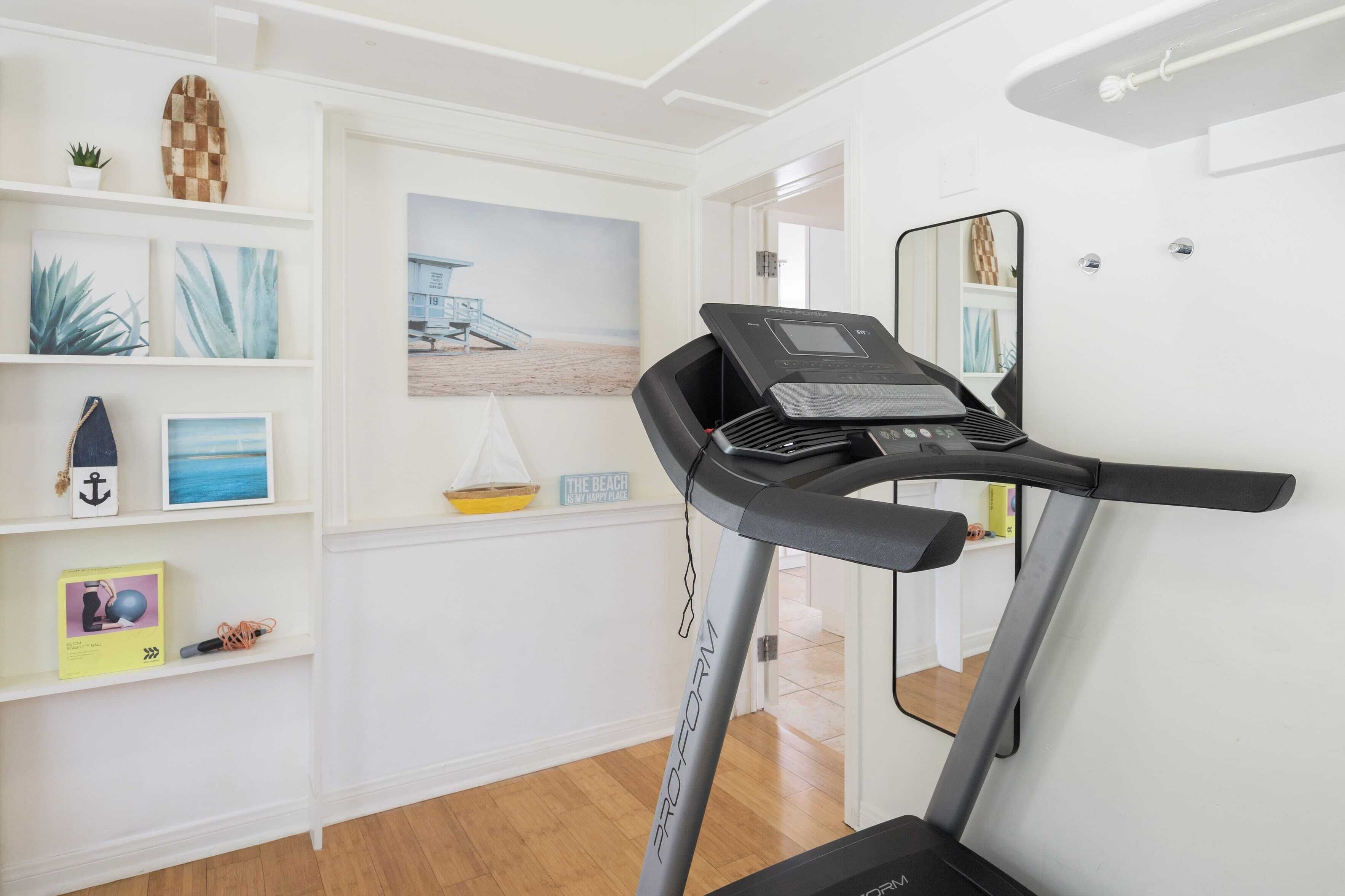 Une salle de sport à domicile équipée d’un tapis de course, d’une étagère avec des objets décoratifs et d’un tableau sur le thème de la plage.
