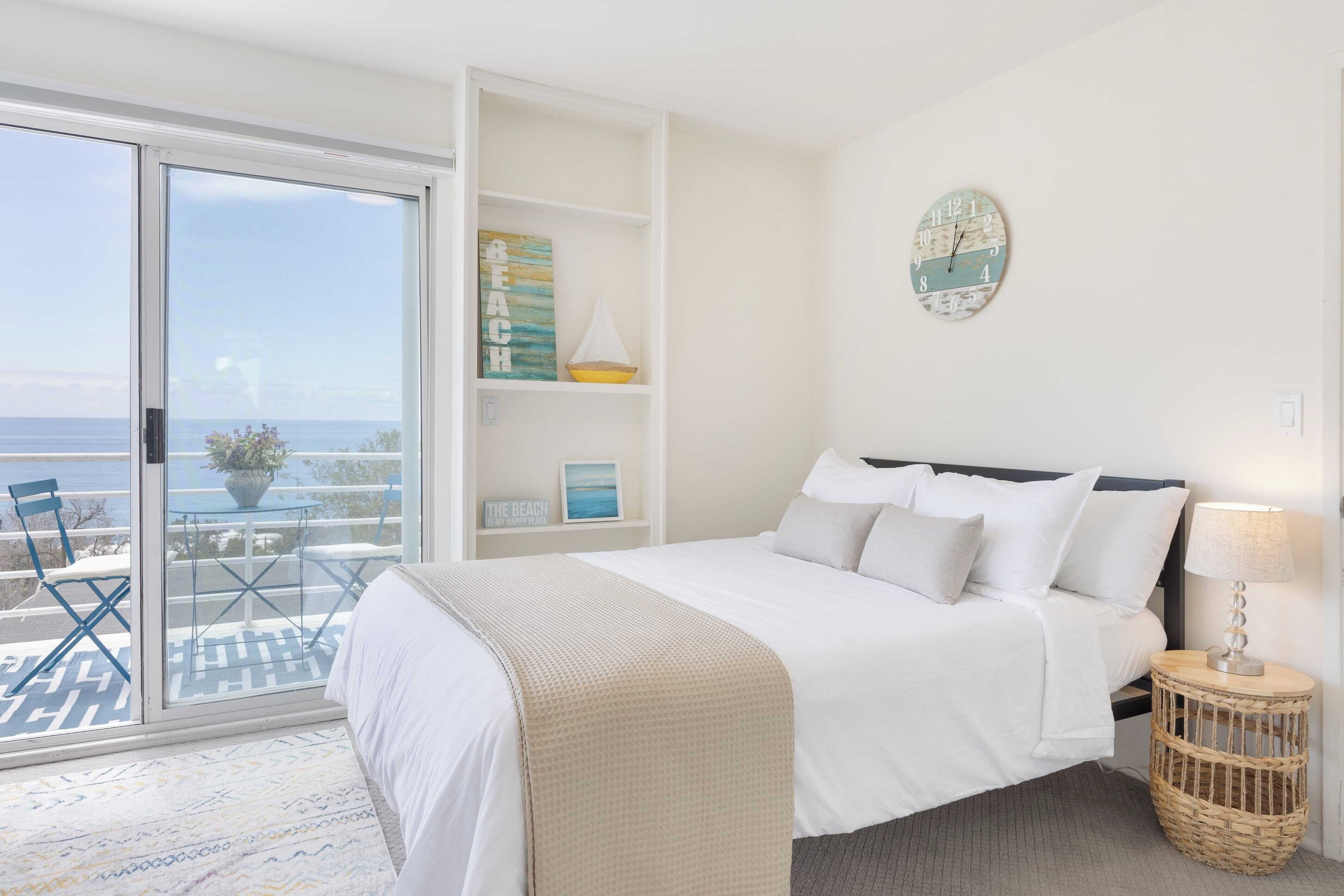 Une chambre à coucher avec un lit, une lampe de chevet, une horloge murale et une vue sur la mer, visible à travers une porte coulissante.