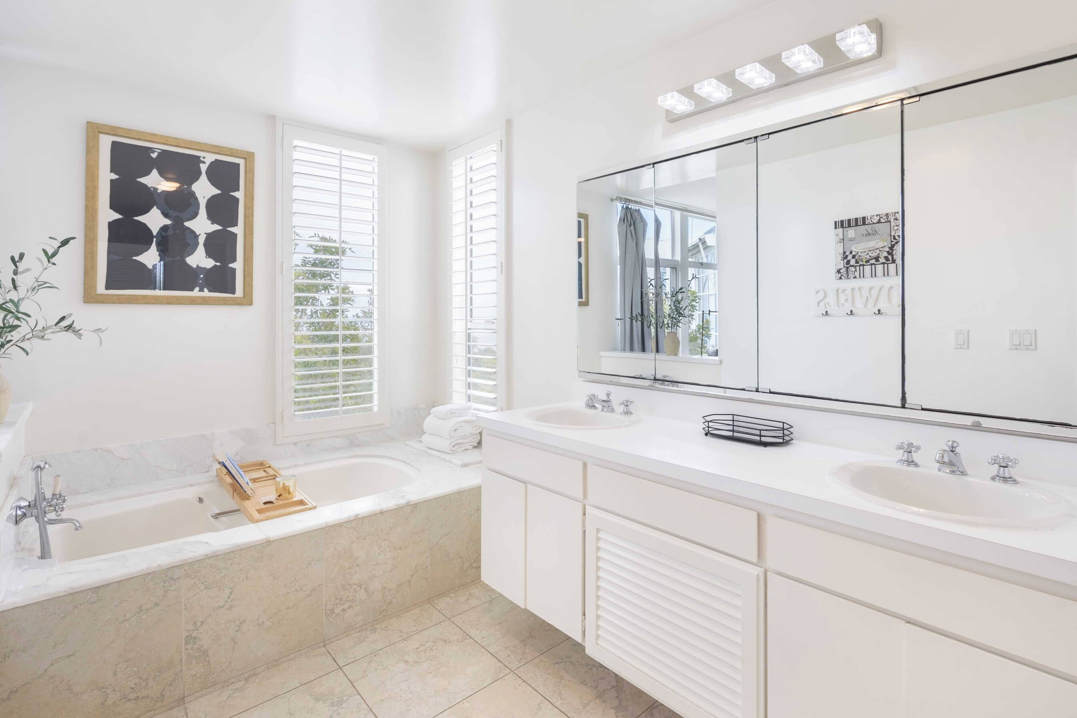 Une salle de bain moderne avec un grand miroir, deux lavabos, une baignoire et une fenêtre équipée de stores.