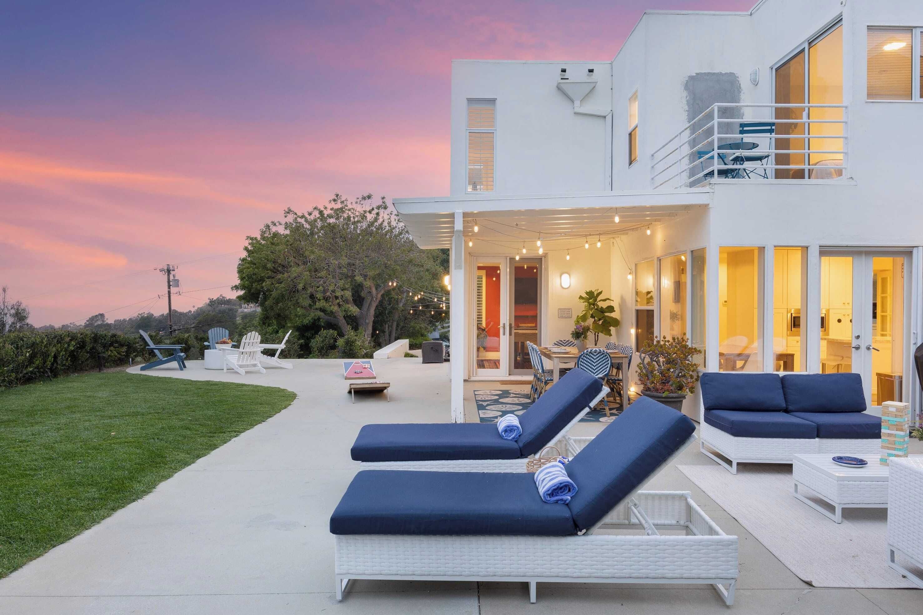 A modern two-story house with a covered patio area featuring lounge chairs and a dining table.