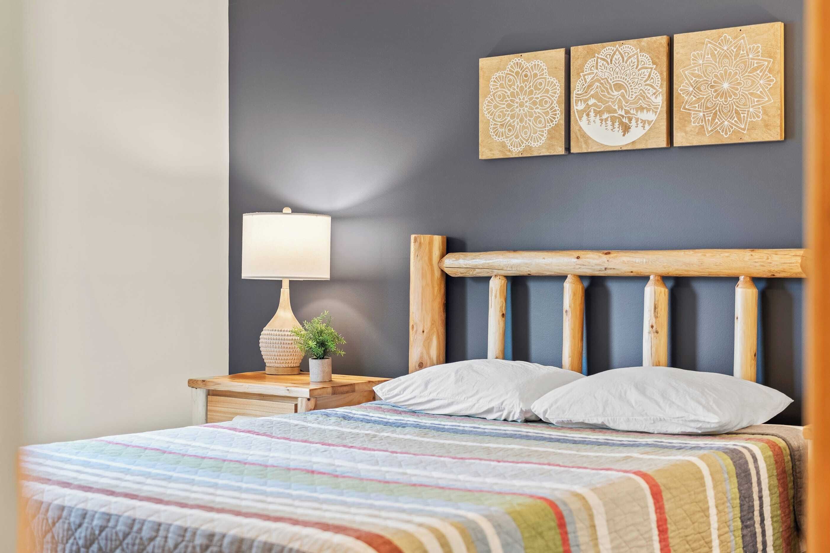 Ein ordentlich bezogenes Bett mit einer bunten, gestreiften Decke, einem Holzkopfschild und einem Nachttisch mit Lampe und einer kleinen Pflanze.