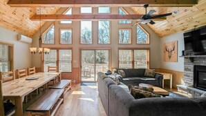 A spacious room with a wooden ceiling, large windows, a fireplace, a sofa, and a dining area. - Bluejay Getaway by Avantstay Private Lake Cabin Hot Tub + Game Room Walk to Lake (Albrightsville)