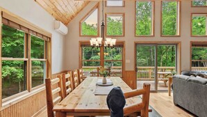 A wooden dining table with chairs, a chandelier, and a view of the forest through large windows. - Bluejay Getaway by Avantstay Private Lake Cabin Hot Tub + Game Room Walk to Lake (Albrightsville)