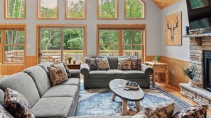 A cozy living room with a gray sofa, patterned cushions, a round coffee table, and large windows offering a view of the outdoors. - Bluejay Getaway by Avantstay Private Lake Cabin Hot Tub + Game Room Walk to Lake (Albrightsville)