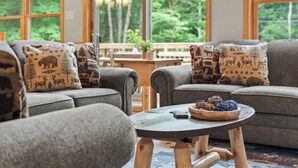 A cozy living area with two grey sofas, each with patterned cushions, a small wooden table with a bowl of fruit, and large windows offering a view of a forested area. - Bluejay Getaway by Avantstay Private Lake Cabin Hot Tub + Game Room Walk to Lake (Albrightsville)
