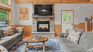 A cozy living room with a stone fireplace, a flat-screen TV, and two grey sofas with patterned cushions.