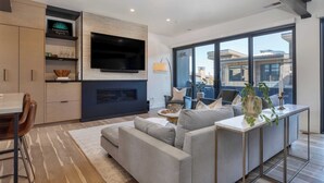 A modern living room with a grey sofa, a TV mounted on a brick wall, and a large window offering a city view. - The Ridge at Canyons Village by Avantstay Stunning Slope-side Condo w/ Hot Tub (Park City)