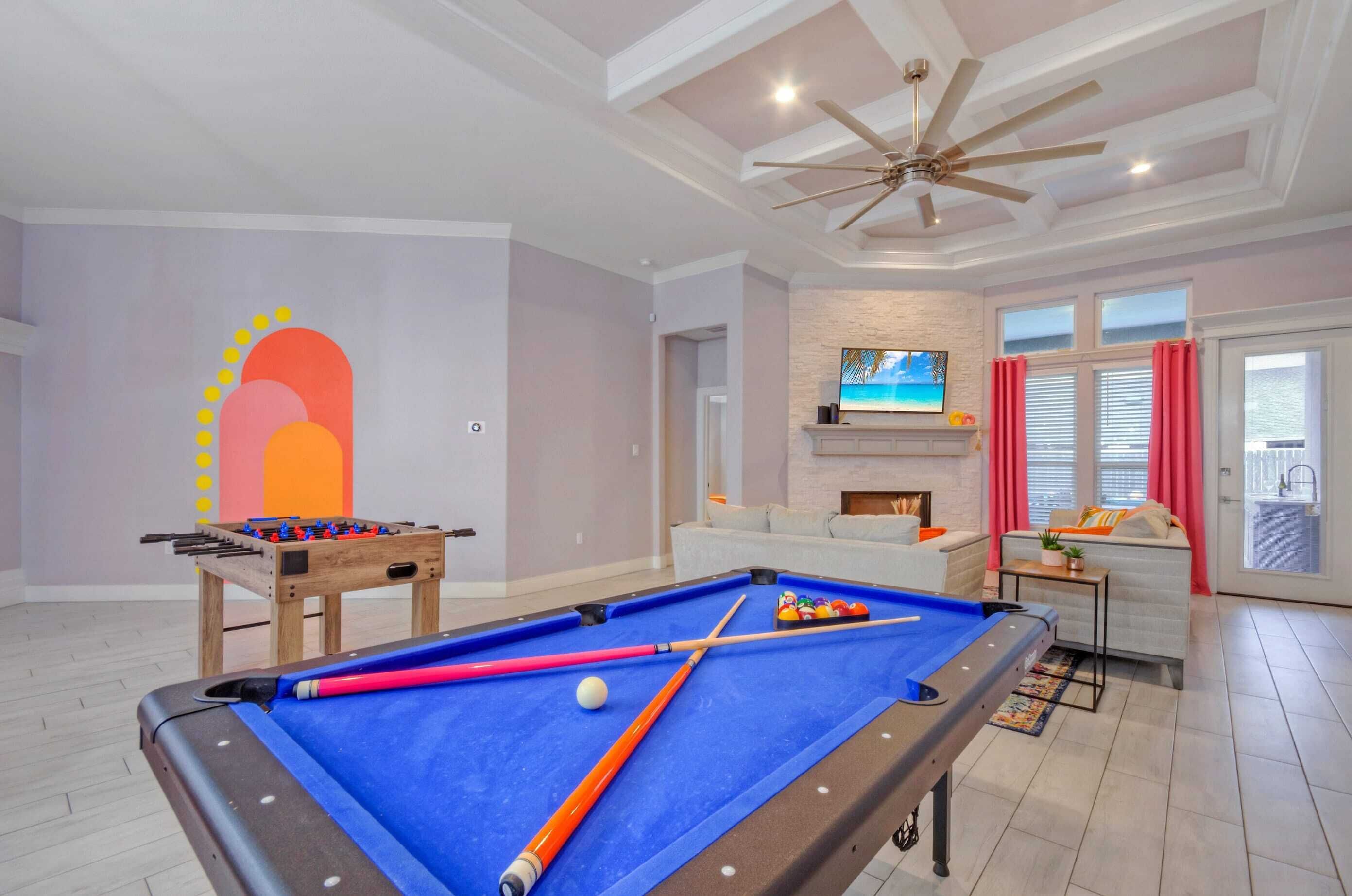 Une table de billard recouverte d’un drap bleu, une table de baby-foot et un téléviseur fixé au mur.