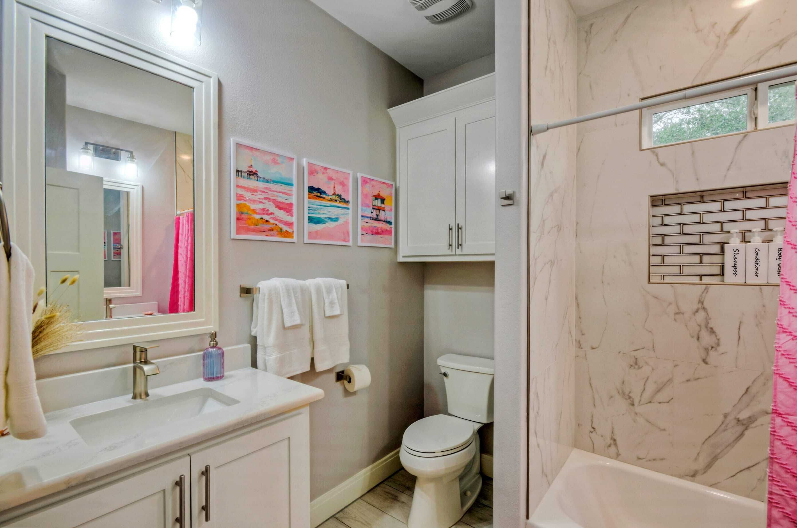 A bathroom with a white sink, mirror, and toilet. There are three colorful paintings on the wall, a white cabinet, and a shower with a marble wall.