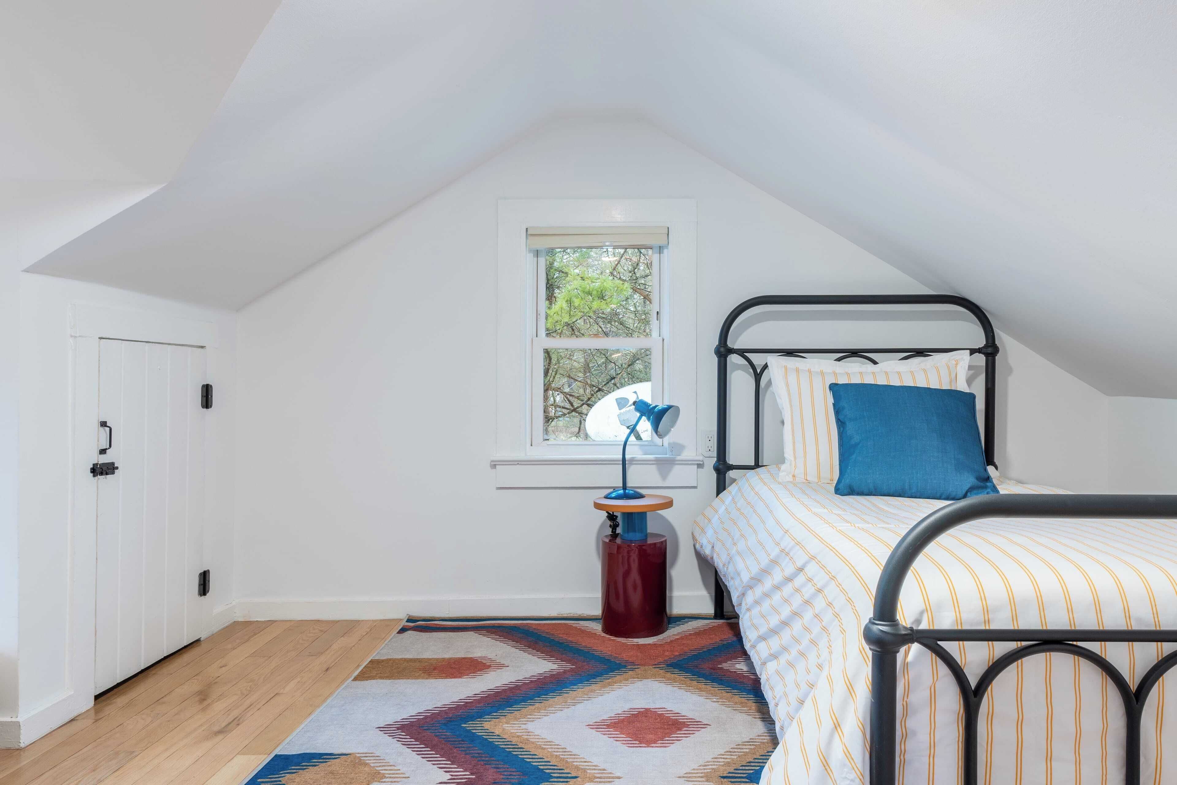 A bedroom with a sloped ceiling, a bed with striped bedding, a blue pillow, a small table with a lamp, and a window with a view of trees.