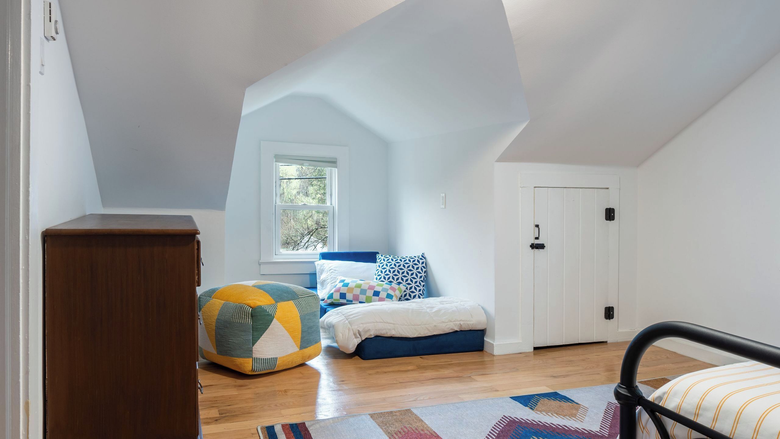 Un dormitorio con techo inclinado, una ventana, una cama con almohadones coloridos, suelo de madera y una puerta.