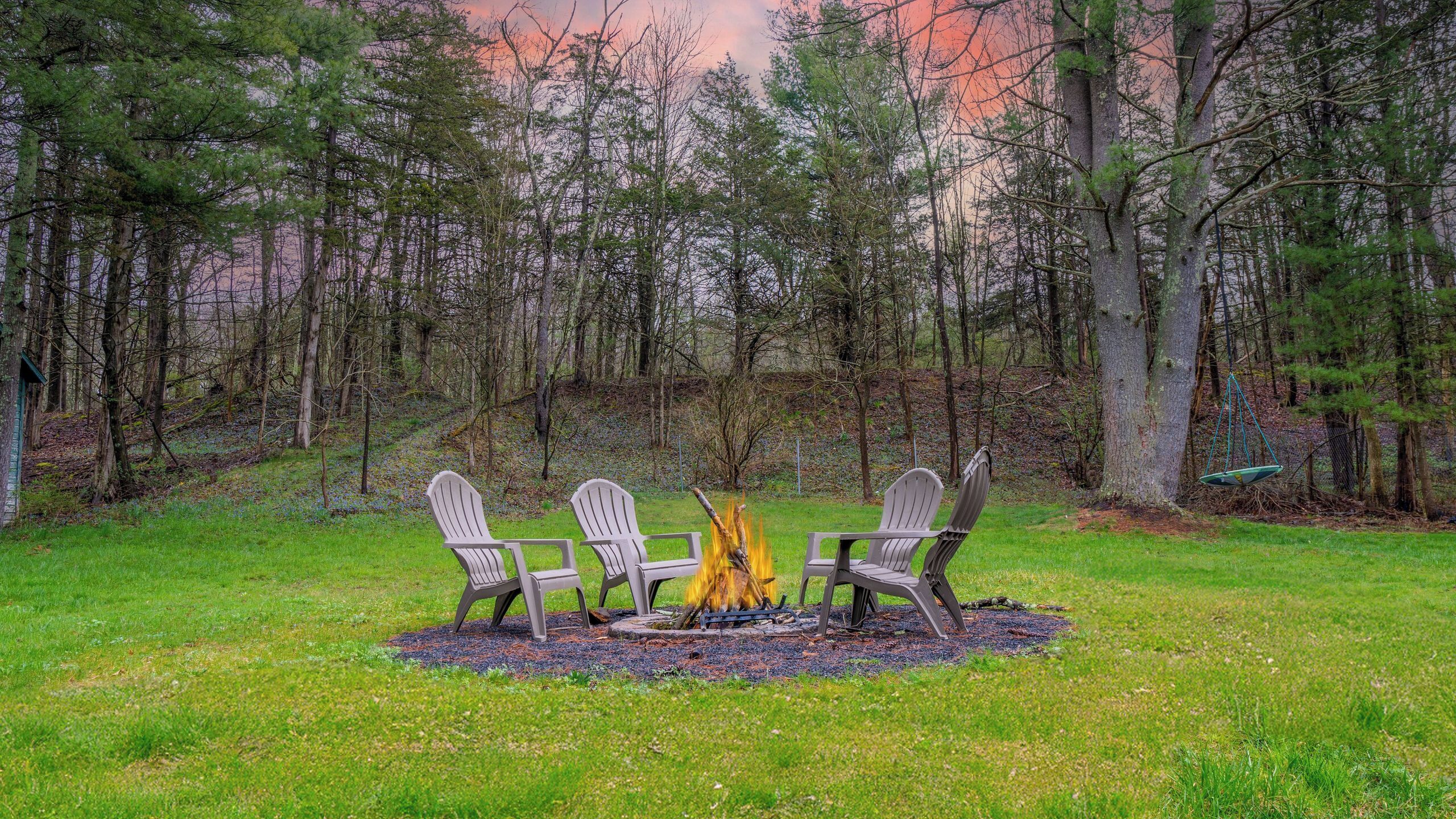 A fire pit with three Adirondack chairs in a forested area.