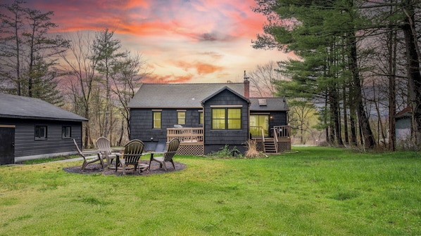A two-story cabin with a deck, surrounded by a grassy yard and trees.