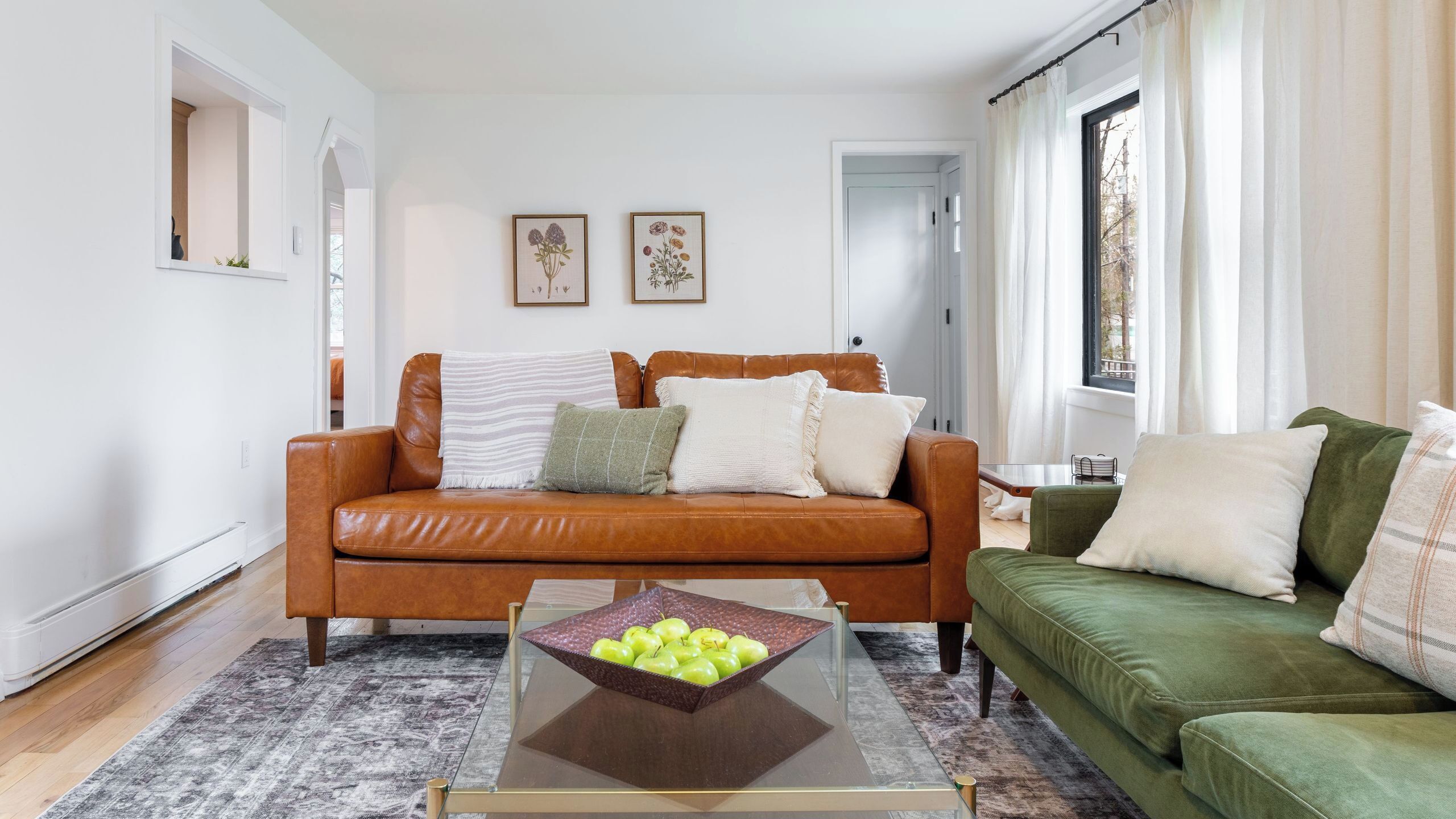 A living room with a brown leather sofa, a green sofa, a coffee table with apples, and two framed floral paintings on the wall.