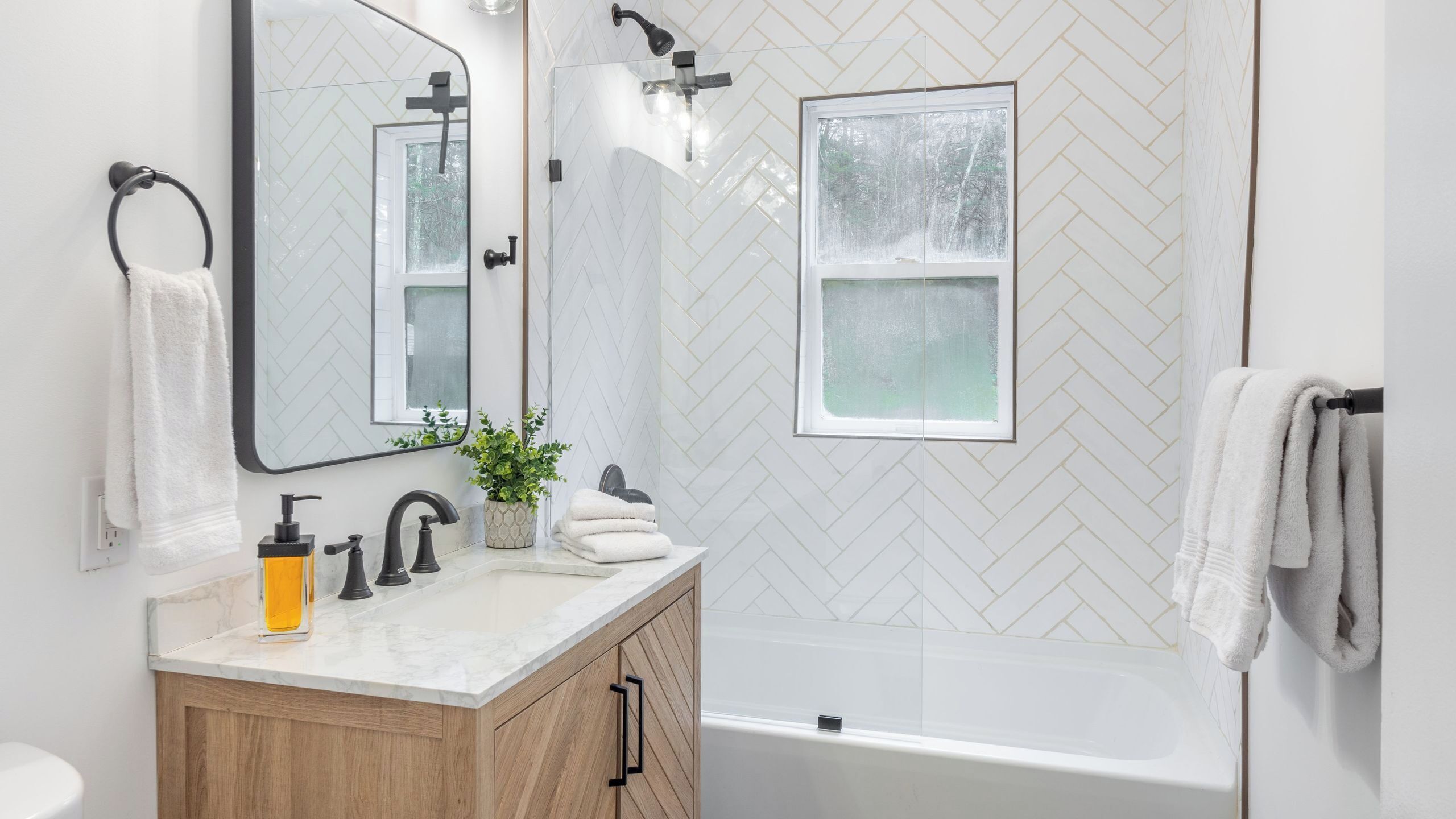 Casa de banho com parede em azulejo branco em padrão herringbone, banheira, pia com espelho e janela.