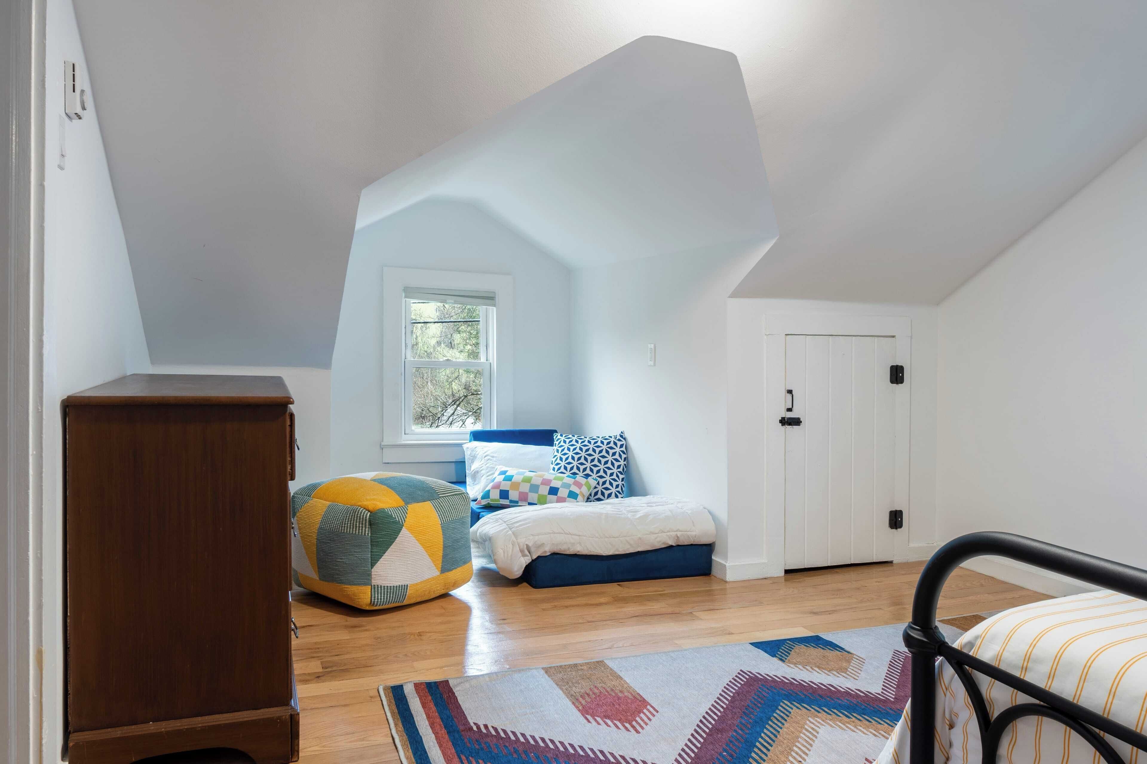 A bedroom with a sloped ceiling, a bed with blue bedding, a colorful bean bag, a wooden cabinet, and a patterned rug.