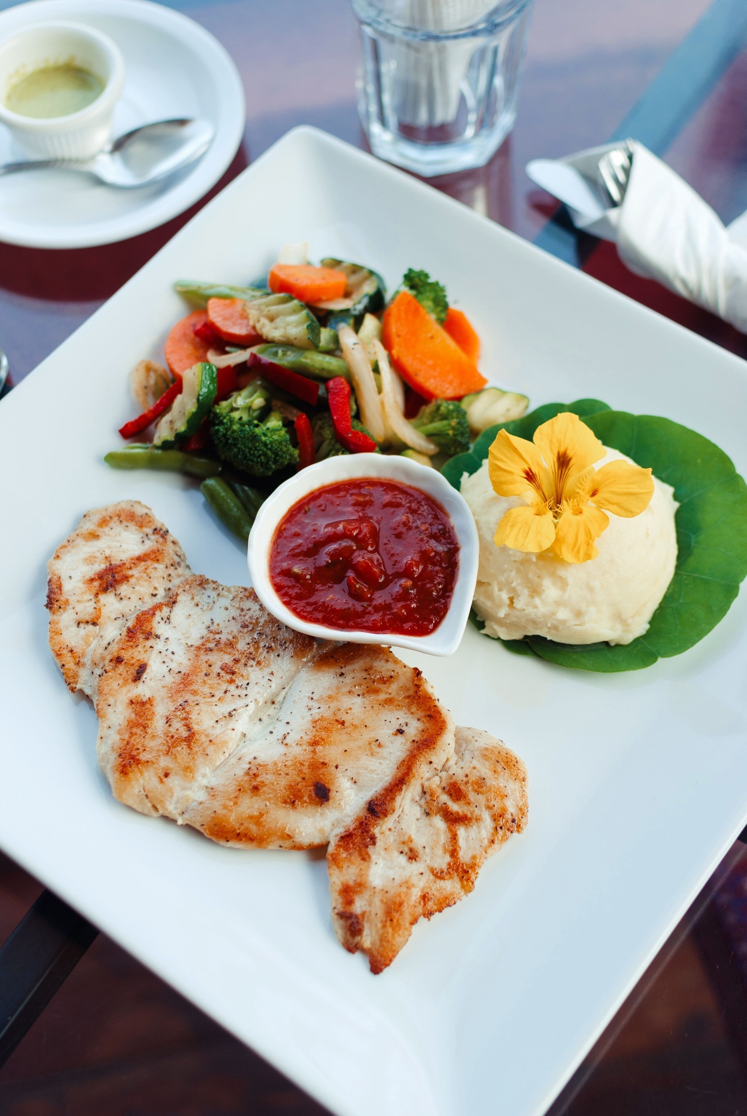 Une assiette blanche contenant du poulet grillé, une salade en accompagnement, de la purée de pommes de terre et une fleur en guise de décoration.